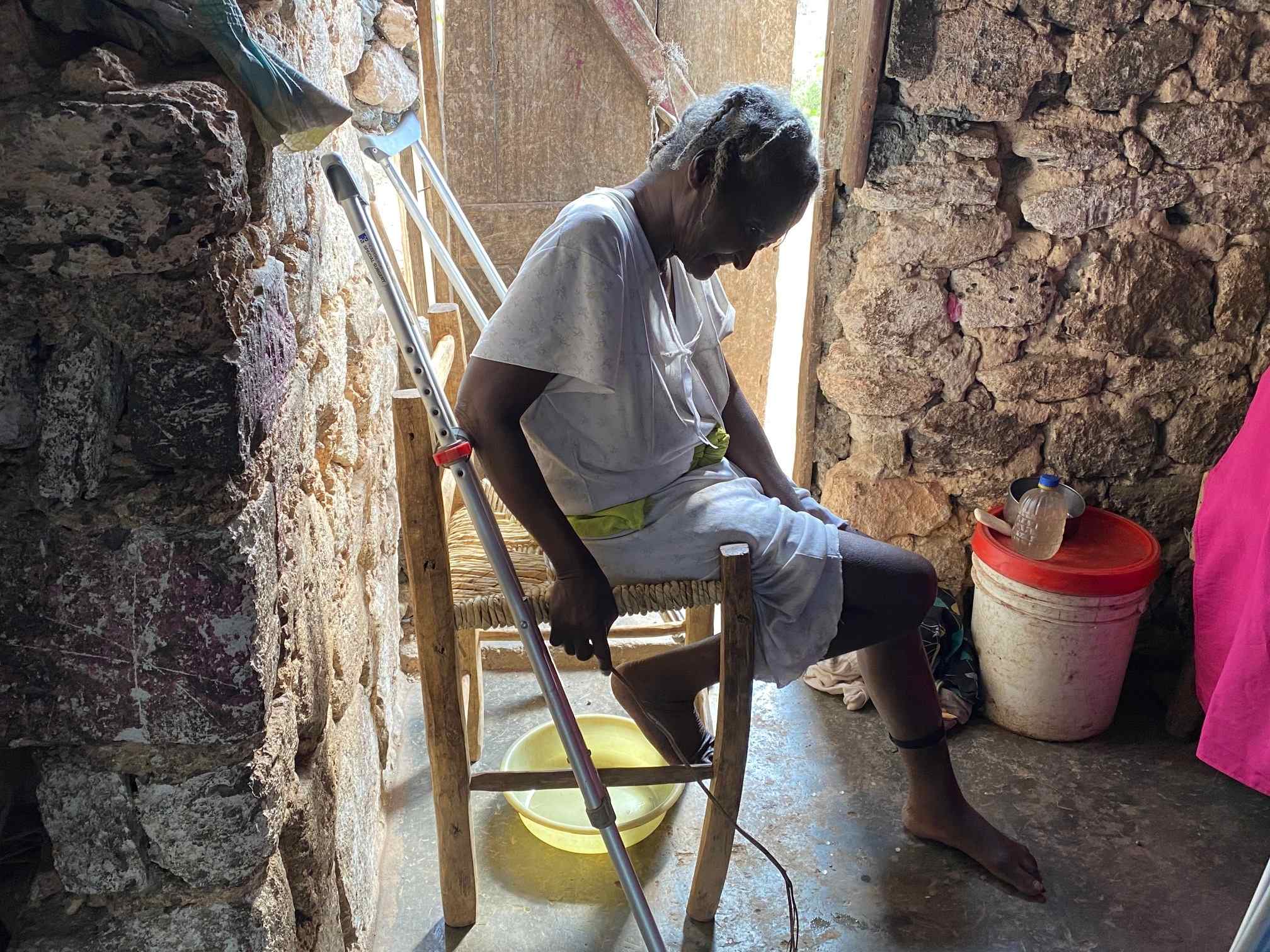 Madame Manie sitting in her stone home in Haiti with her crutches nearby, illustrating a 'Love in Action' visit from Many Hands' Spiritual Shepherds providing food and prayer for her family.