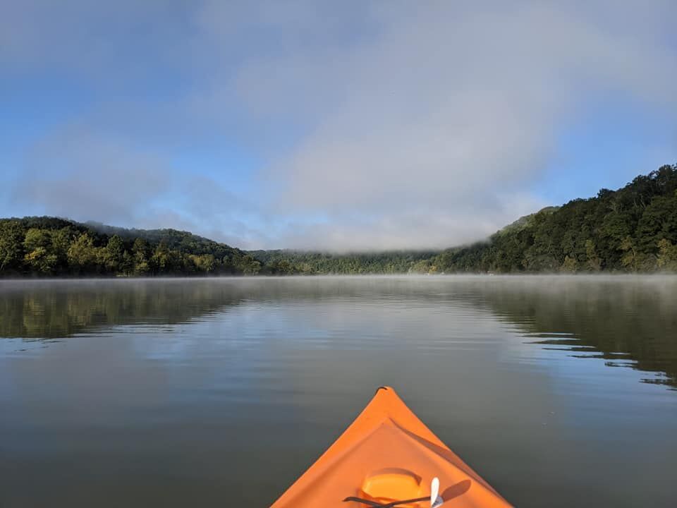 The bow of an orange kayak gliding across a calm, misty lake at dawn, symbolizing the quiet moments of reflection and vision-casting for Many Hands' future initiatives.