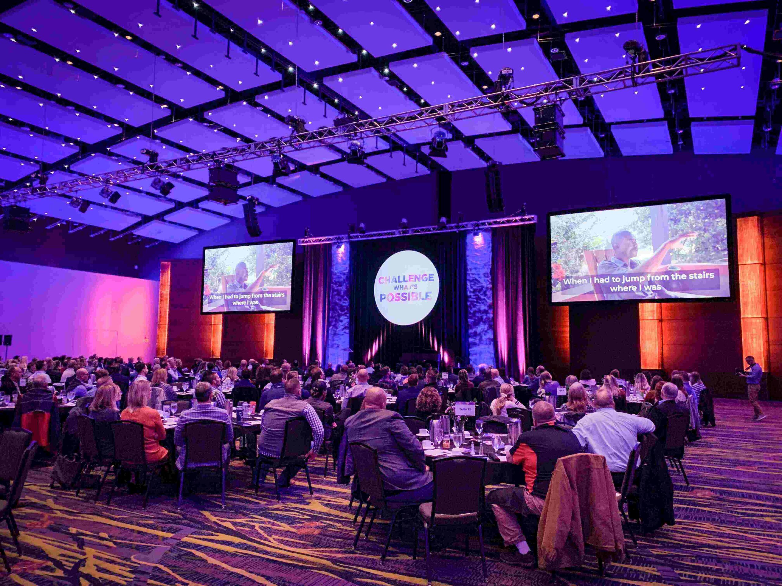 A large crowd of over 250 attendees at the third annual Many Hands Business Breakfast in a professional ballroom, featuring keynote screens and the theme 'Challenge What’s Possible.