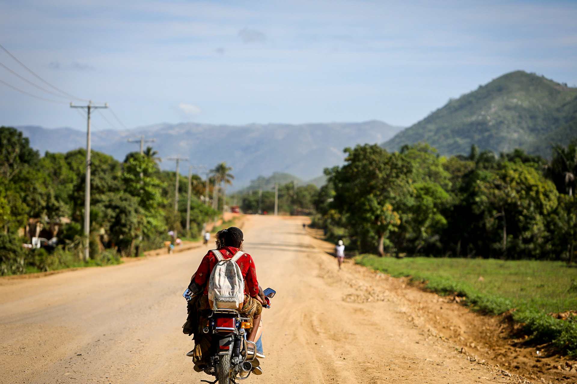 A person on a motorcycle traveling down a long, rural dirt road in Sylvain, Haiti, representing the humble beginnings and the 'path' where the Many Hands Business Breakfast was first dreamed.