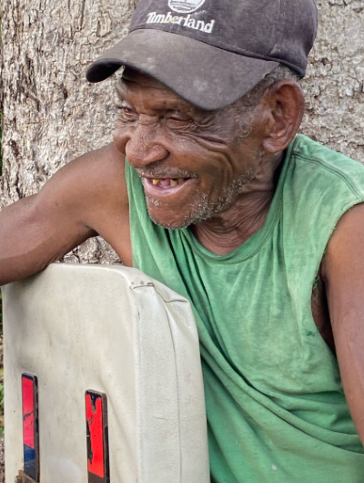 Letwa, an older man in a green tank top and a brown Timberland cap, laughing and looking off-camera while leaning on a white chair in front of a large tree.