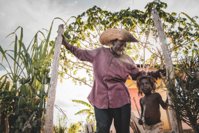 An elderly man in a straw hat shares a joyful moment with a young child in a rural garden setting—highlighting Many Hand’s commitment to community welfare and intergenerational support in Haiti.