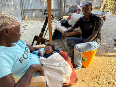 An older woman cradling a sleeping newborn in an outdoor setting, with a man sitting opposite them on a container, representing Many Hands' community-based support network for mothers and infants in Haiti.