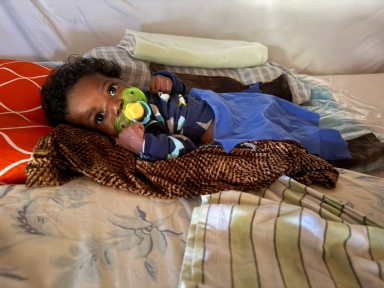 A close-up photograph of a newborn baby lying on a makeshift bed, using a pacifier and gazing intently at the camera, illustrating Many Hands' focus on maternal health and early childhood survival programs in rural Haiti.