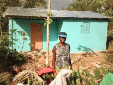 A woman stands in front of a turquoise blue home with a silver tin roof, built through the Many Hands Homes for Haiti Hallelujah (HHH) legacy project.