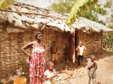 A Haitian mother and her children stand before their traditional woven-branch home with a thatched roof, illustrating the need for sustainable housing outcomes in Pignon.