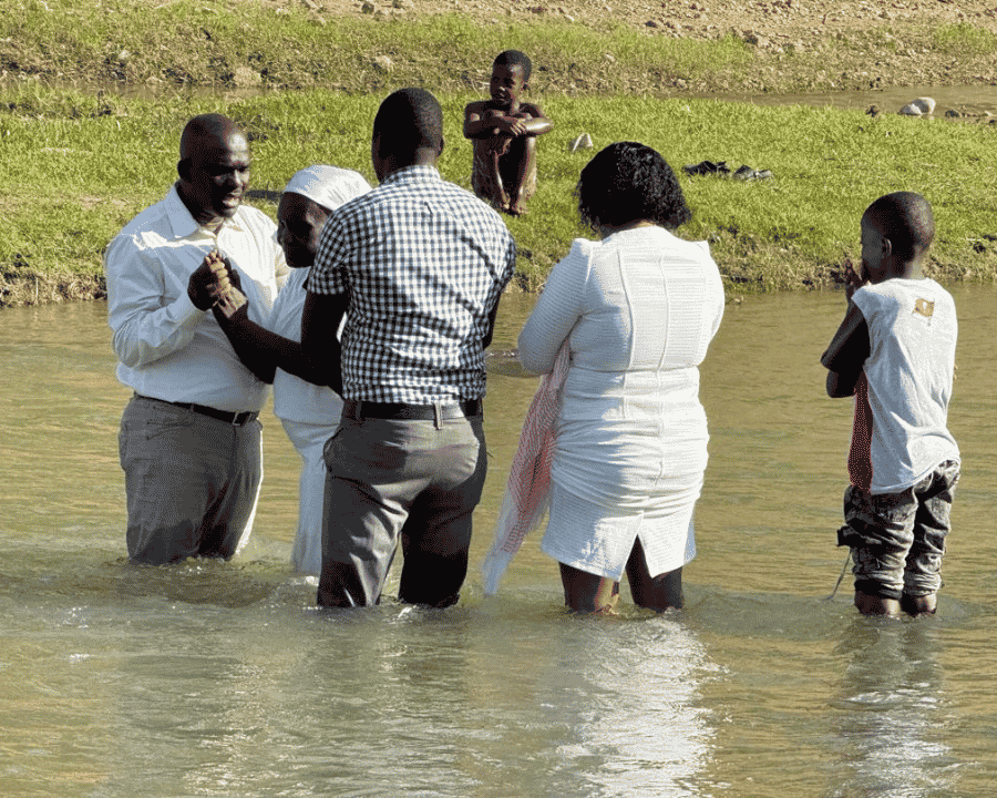 Louisa, at age 69, stands in the Bouyara river surrounded by her local pastor and community members during her Christian baptism, representing a powerful spiritual breakthrough in her faith journey.