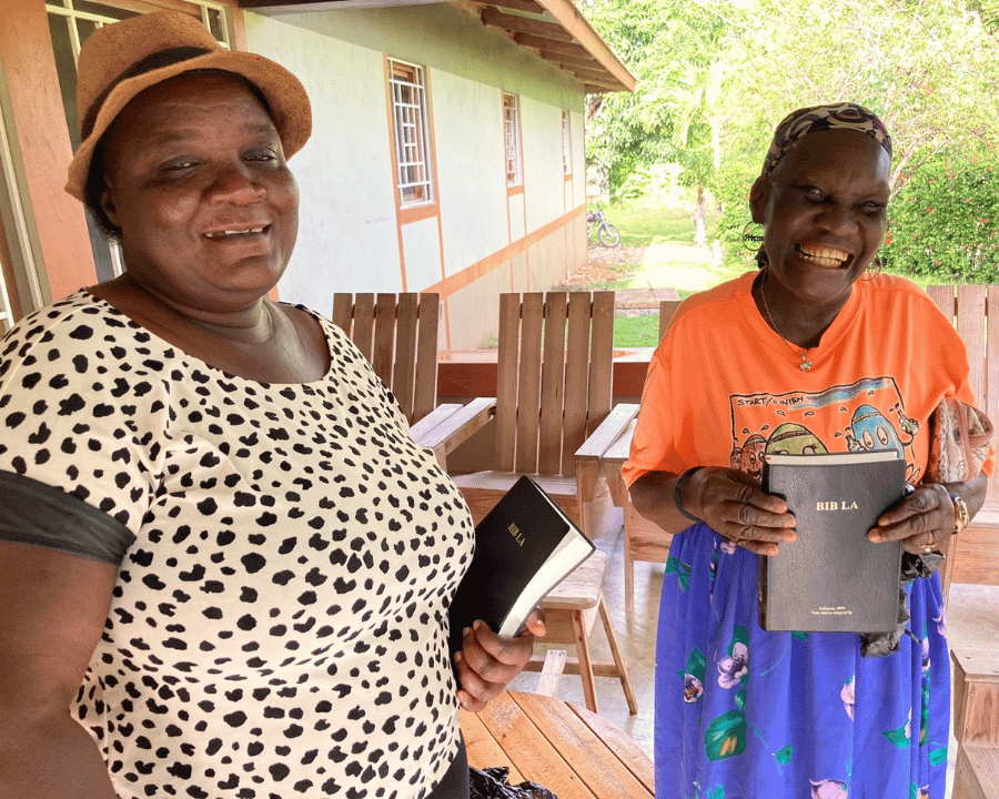 Many Hands staff member Yranna stands proudly alongside Louisa as they smile and hold their new Creole Bibles, illustrating the power of relational discipleship and holistic transformation in Sylvain, Haiti.