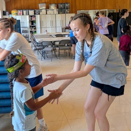 A DMC youth volunteer bends down to play a handclapping game with a young child wearing a paper crown, embodying the mutual joy and connection of an Abaco impact trip.