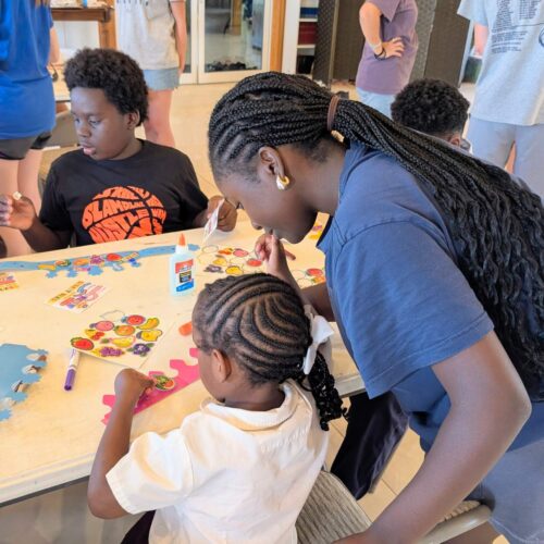 A DMC team member leans over to help a young Bahamian girl with an art project during a church kids camp, highlighting Many Hands' focus on relational ministry and life development.