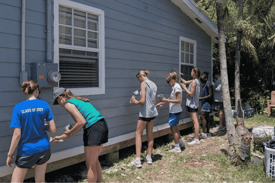 Volunteers from the Des Moines Christian School impact team line up to paint the exterior siding of a local home, supporting Many Hands' Safe Structures housing initiatives in Abaco.