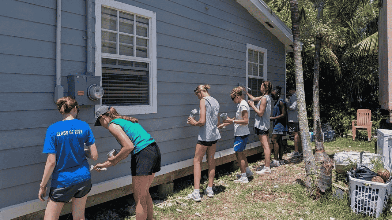 Volunteers from the Des Moines Christian School impact team line up to paint the exterior siding of a local home, supporting Many Hands' Safe Structures housing initiatives in Abaco.