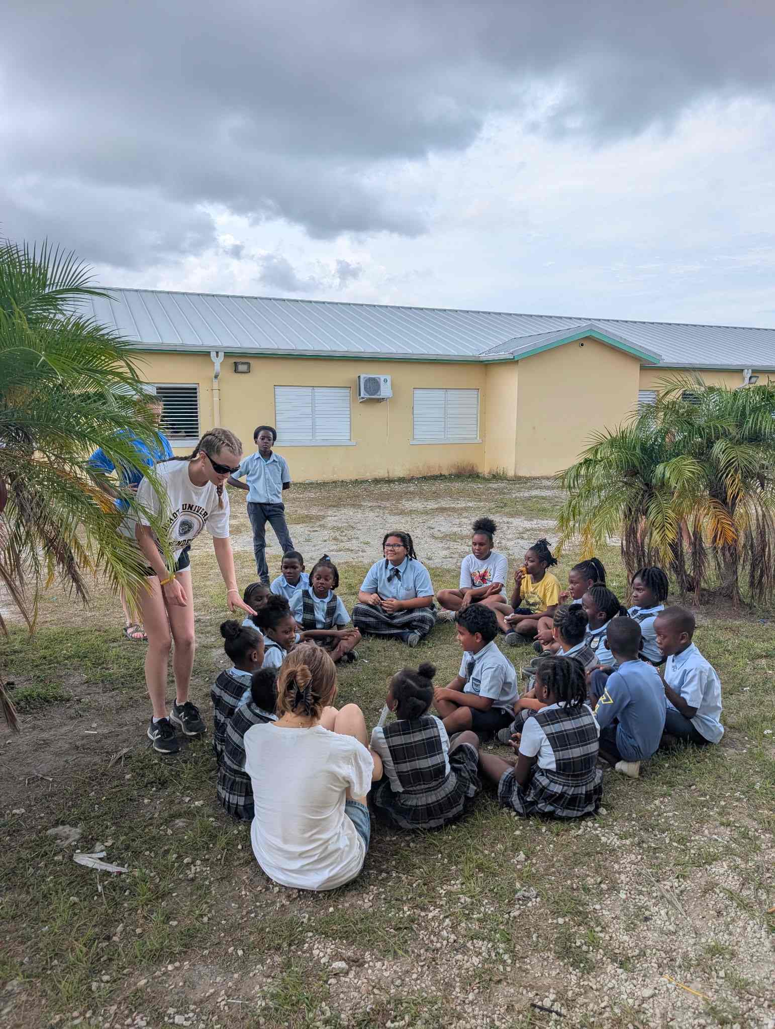 A DMC impact trip volunteer bends forward to interact with a circle of local Bahamian students seated on the grass during recess at a public primary school.