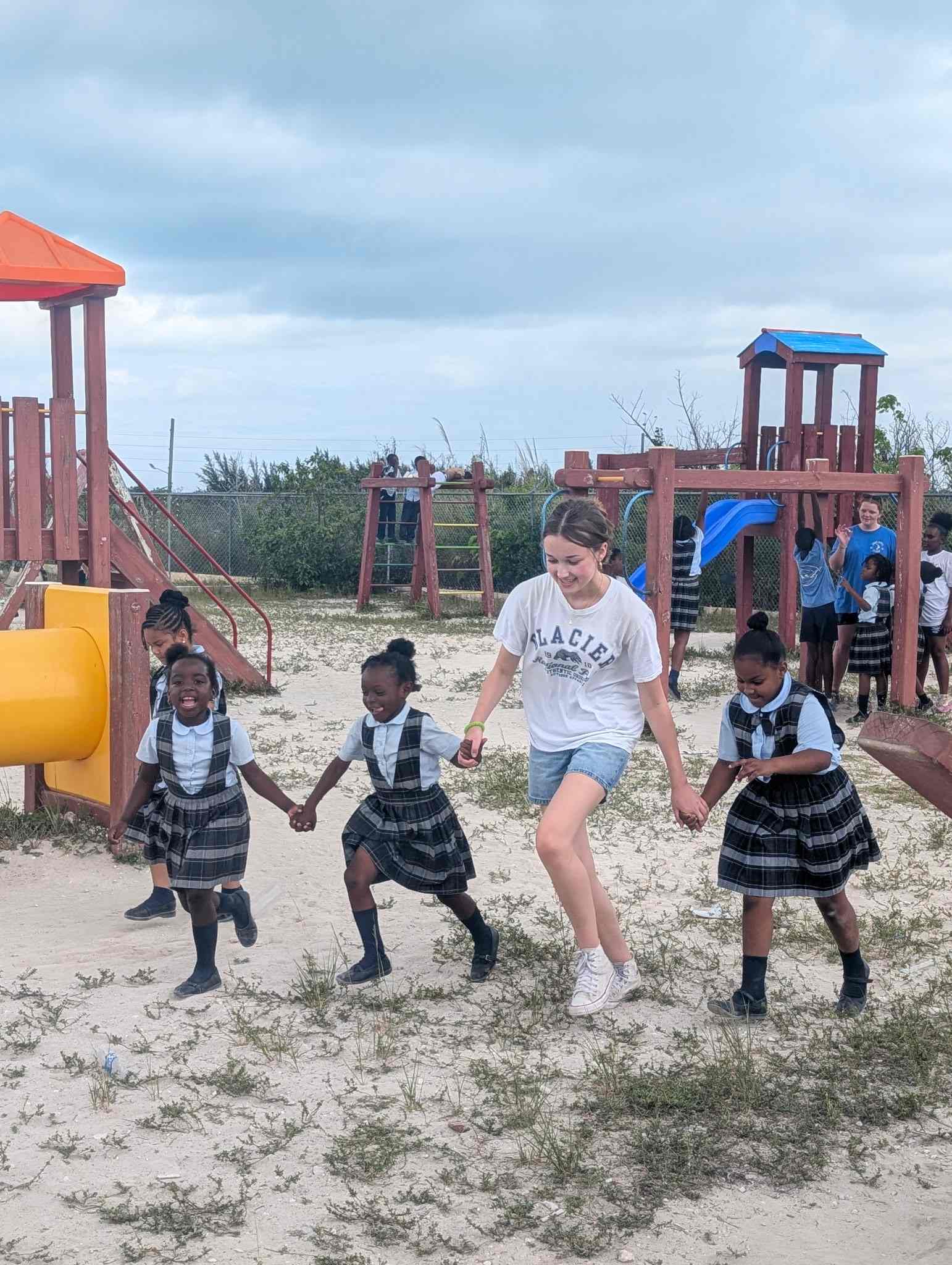 A teenage student volunteer holds hands and runs across the playground with local primary students during a school recess visit focused on community connection.