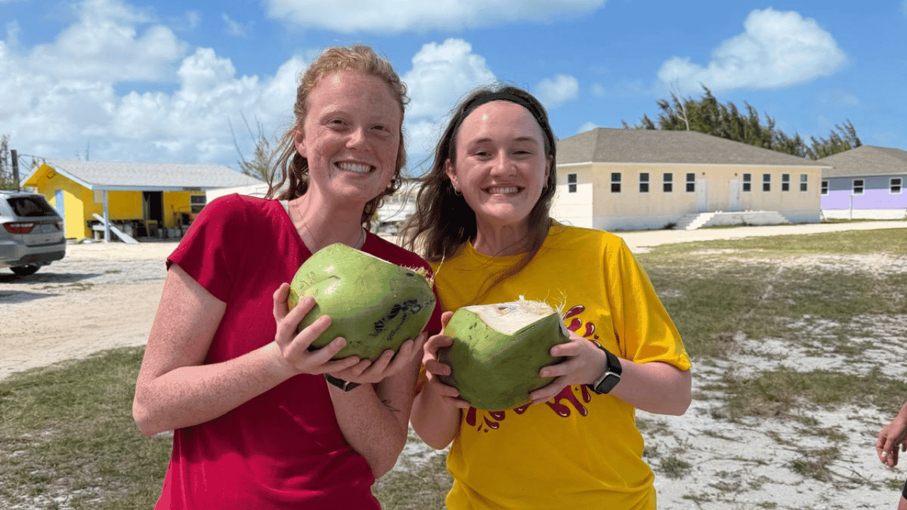 Two female volunteers from Lutheran Church of Hope Ames smile while holding fresh green coconuts, experiencing local hospitality and Bahamian culture on a Many Hands trip.