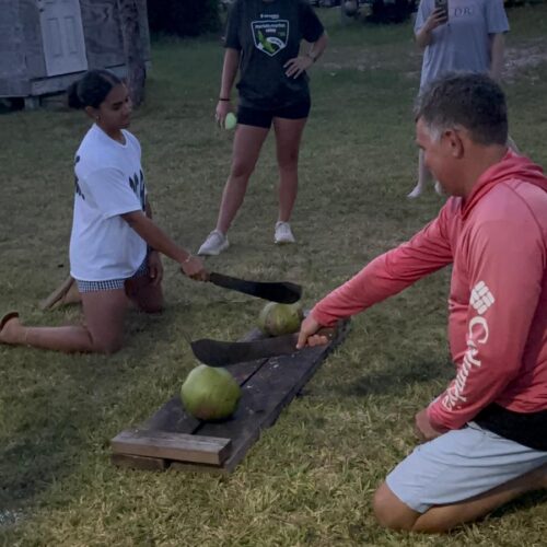 Volunteers and local leaders use machetes to split fresh coconuts during a final evening team gathering, demonstrating the welcoming hospitality and cultural immersion of an Abaco impact trip.