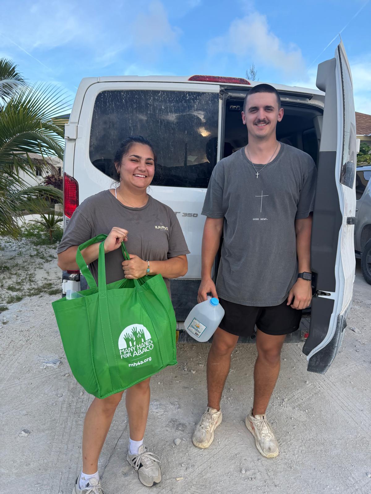 Two Kairos impact team members hold a green 'Many Hands for Abaco' food distribution bag and water jug while delivering essential supplies to local families.