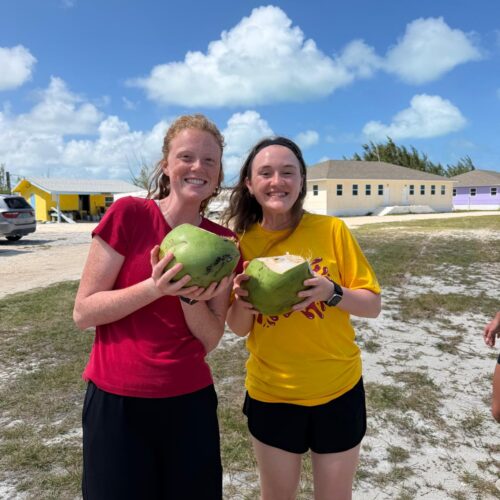Two female volunteers from Lutheran Church of Hope Ames smile while holding fresh green coconuts, experiencing local hospitality and Bahamian culture on a Many Hands trip.