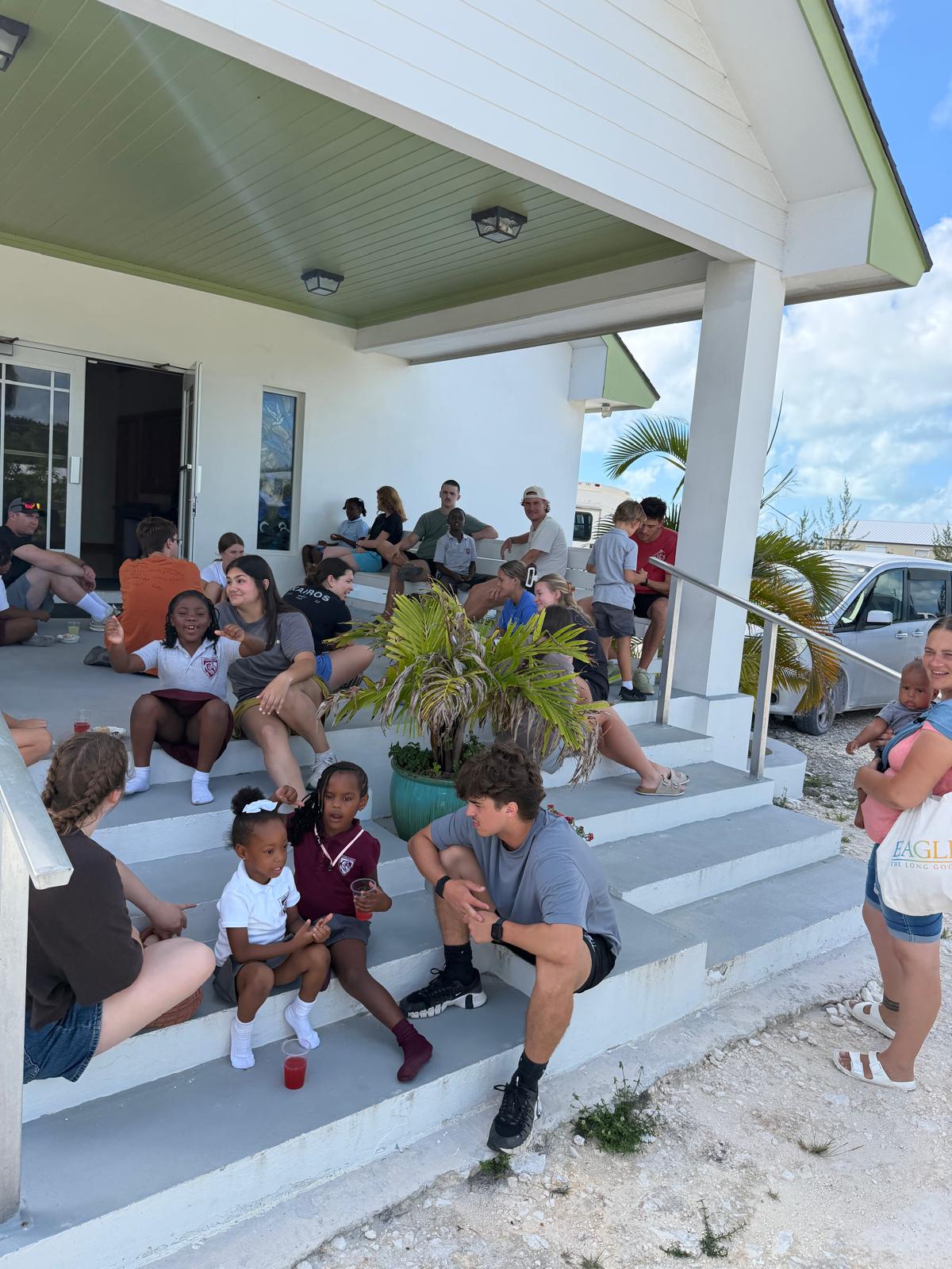 Many Hands volunteers and local kids sit together on church steps, showcasing community connection and life development programs following storm recovery efforts in Abaco.