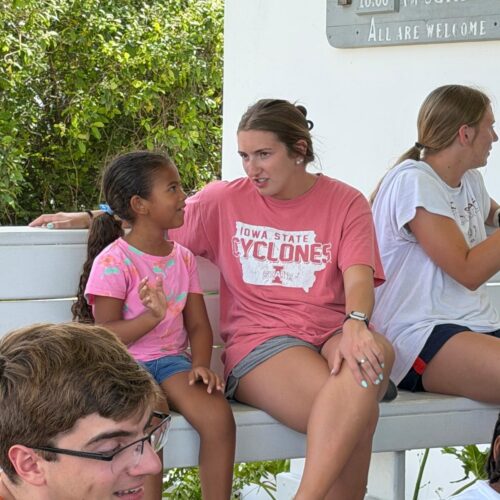 An Iowa State University student volunteer chats warmly with a young girl on a bench, embodying Many Hands' mission to honor individual stories and build deep community trust.