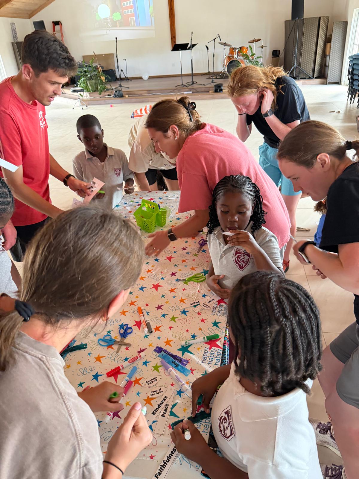 Many Hands Kairos team volunteers sit with local children around a colorful table to create crafts during a faith-based summer kids camp in Abaco, Bahamas.
