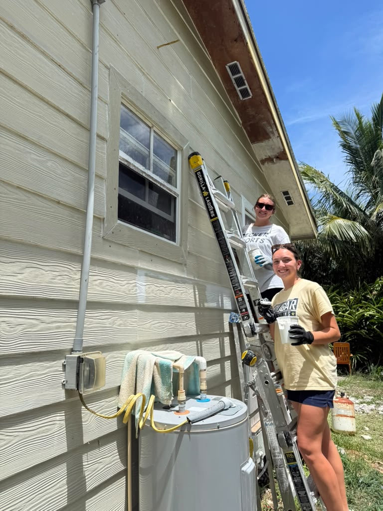 Many Hands volunteers scale a ladder to paint the exterior trim of Davia Dean's house, participating in community-led hurricane recovery efforts in Abaco.