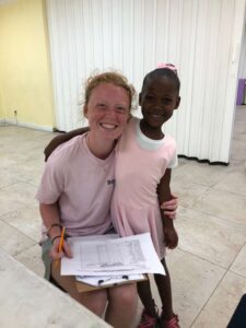A Many Hands Kairos team volunteer smiles next to a young student during a reading mentorship activity at Central Abaco Primary School (CAPS) in the Bahamas.