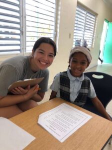 A Many Hands Kairos team volunteer smiles next to a young student during a reading mentorship activity at Central Abaco Primary School (CAPS) in the Bahamas.