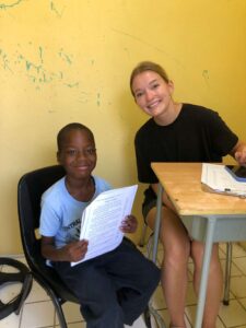 A Many Hands Kairos team volunteer smiles next to a young student during a reading mentorship activity at Central Abaco Primary School (CAPS) in the Bahamas.