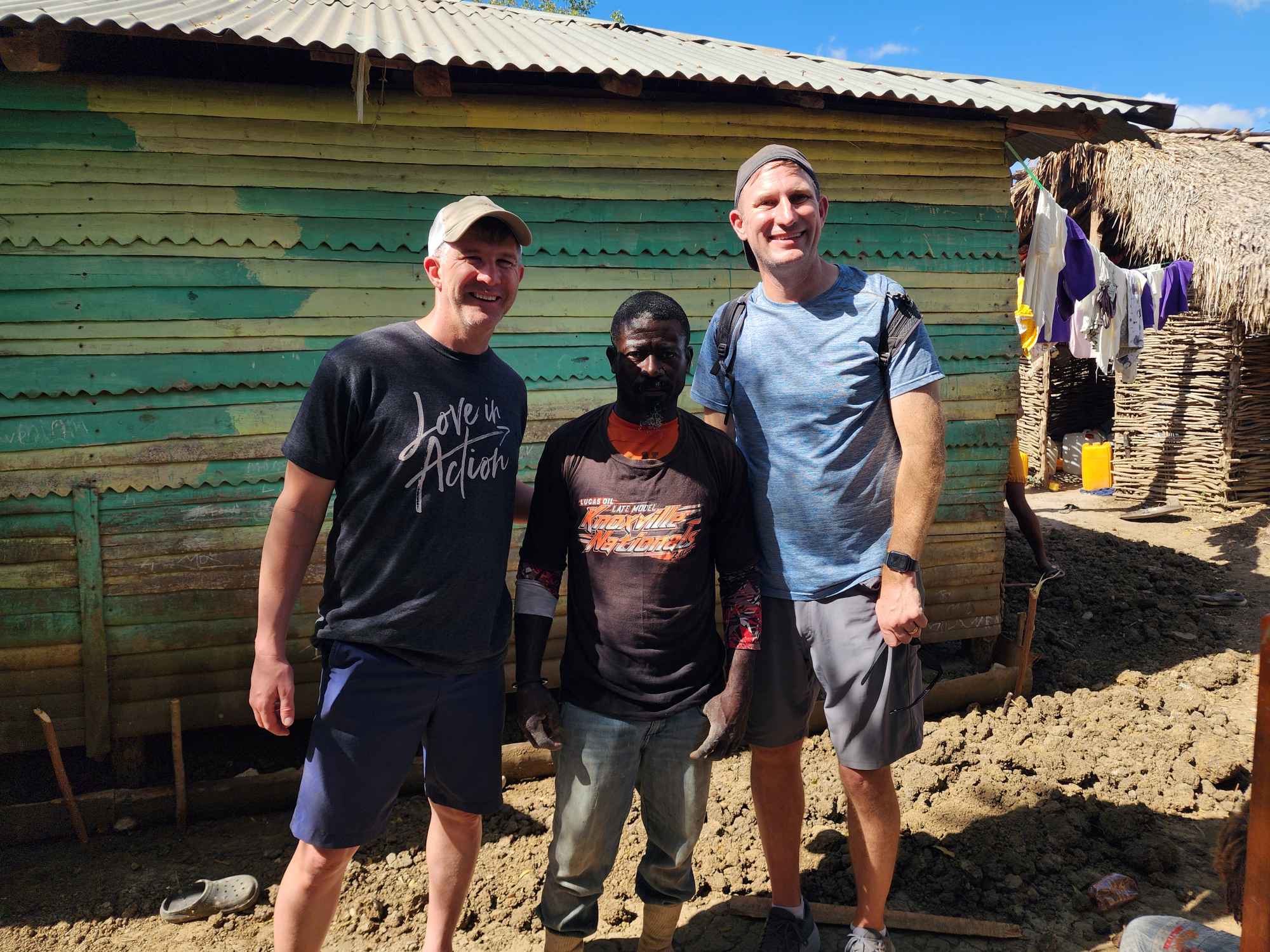 Pastor Andrew Schmidt stands arm-in-arm with a local Haitian resident outside a fragile wooden shelter, illustrating the power of proximity and global compassion.