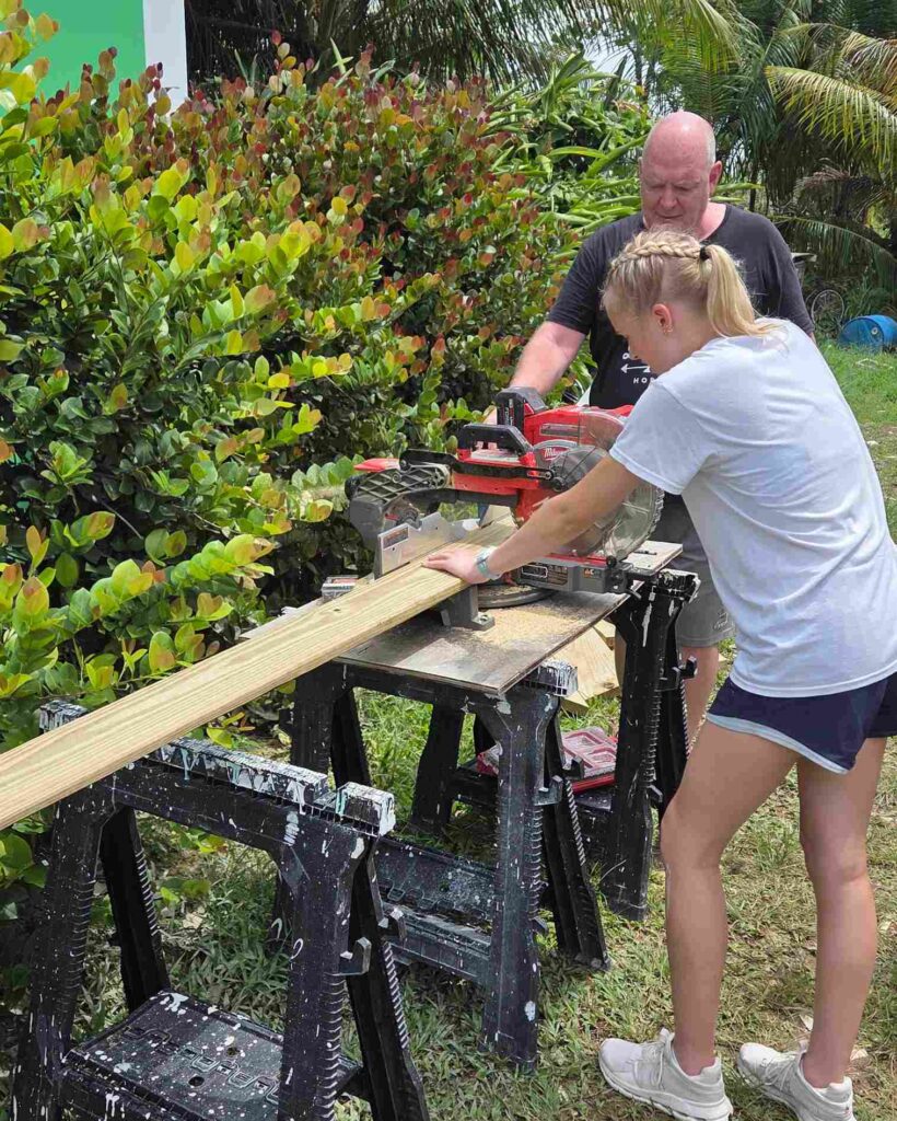 An Immanuel CRC youth volunteer operates a miter saw under adult supervision, gaining hands-on construction experience during a Many Hands home restoration trip in Abaco.