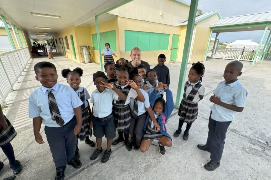 An Immanuel CRC youth volunteer poses with a large group of smiling primary students outside their classrooms during an education support visit to Central Abaco Primary School.