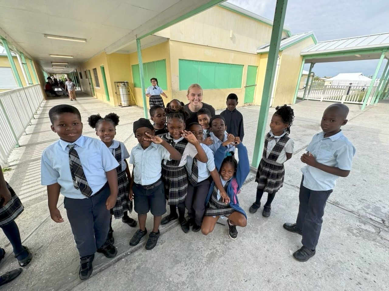 An Immanuel CRC youth volunteer poses with a large group of smiling primary students outside their classrooms during an education support visit to Central Abaco Primary School.