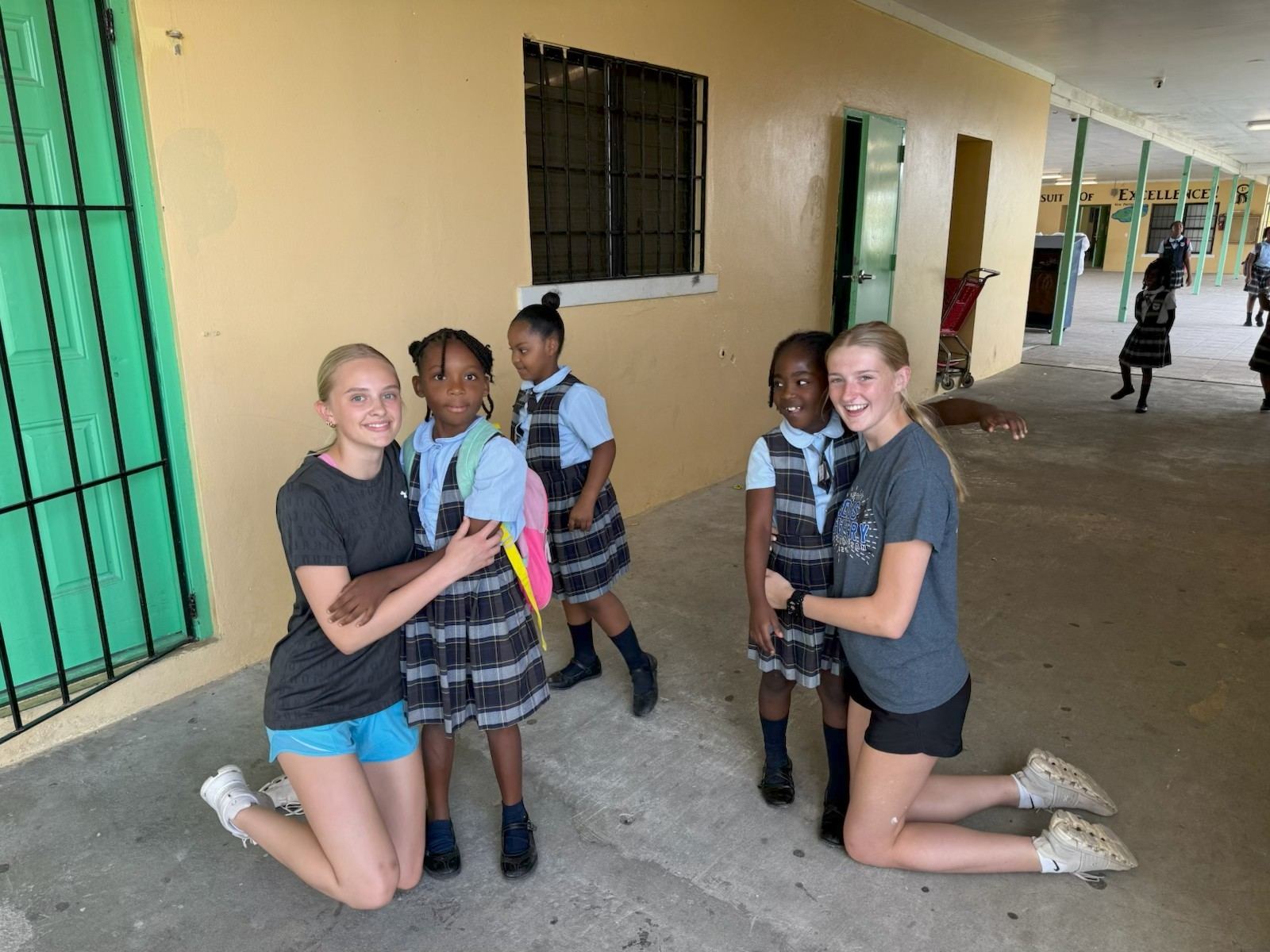 Two female student volunteers kneel down to hug local primary school girls in the hallway, demonstrating the power of personal relationships during an impact trip.