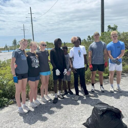 Many Hands volunteers link arm-in-arm with local Bahamian youth during a joint hurricane recovery trash cleanup project at a local high school track field.