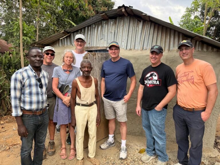 Many Hands team members pose with an elder local resident in front of a fragile mud and corrugated tin shelter, illustrating the core survival needs addressed by our community development projects.