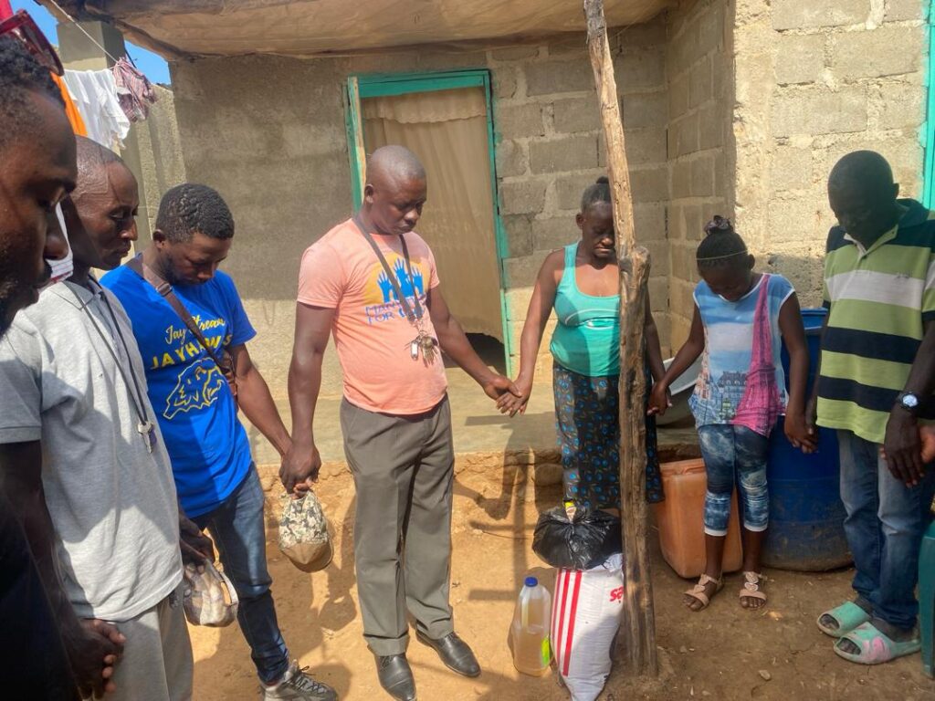 Staff Praying Home Visit 2024 2 Haitian staff members from the Many Hands team hold hands in a prayer circle with a local family during a neighborhood home visit and food supply delivery in Pignon, Haiti.