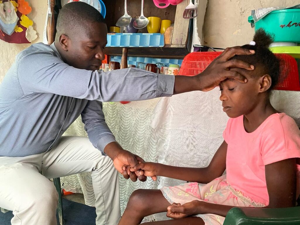 Staff Praying Home Visit 2024 Jean Rupert 2 A local Many Hands staff leader places a hand on a young girl's forehead to pray over her, embodying the deep relational ministry and spiritual care that anchors our field operations.