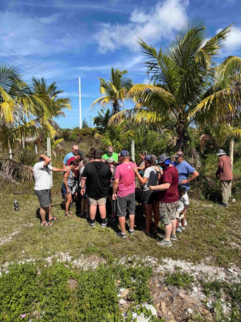 A large huddle of short-term volunteers and local partners form a unified prayer circle under palm trees during a community outreach and distribution ministry day.