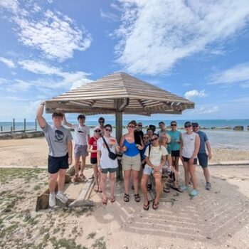 The DMC student impact team gathers for a group photo under a wooden beach shelter, celebrating local culture and collective action during their final days in Abaco.