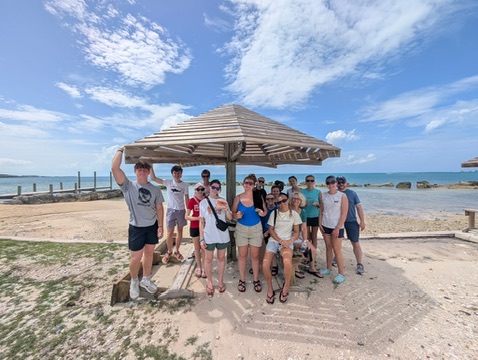 The DMC student impact team gathers for a group photo under a wooden beach shelter, celebrating local culture and collective action during their final days in Abaco.