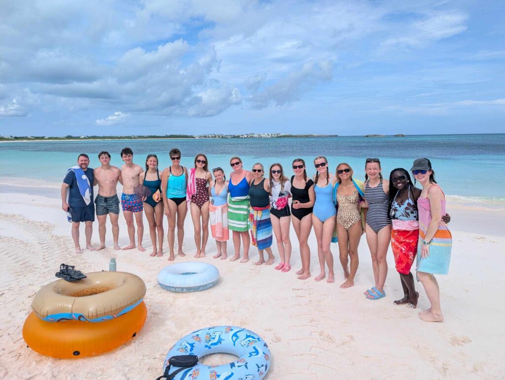 The DMC student impact team stands together on the white sands of Winding Bay, experiencing the natural beauty of the Bahamas on a Many Hands team-building trip.