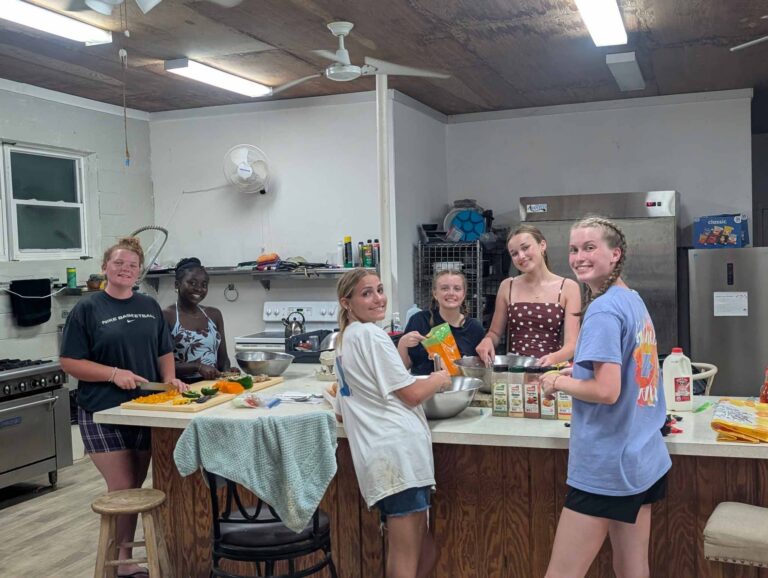 DMC youth volunteers smile together while chopping fresh vegetables and preparing communal meals in the campus kitchen during their Many Hands IMPACT trip to Abaco.