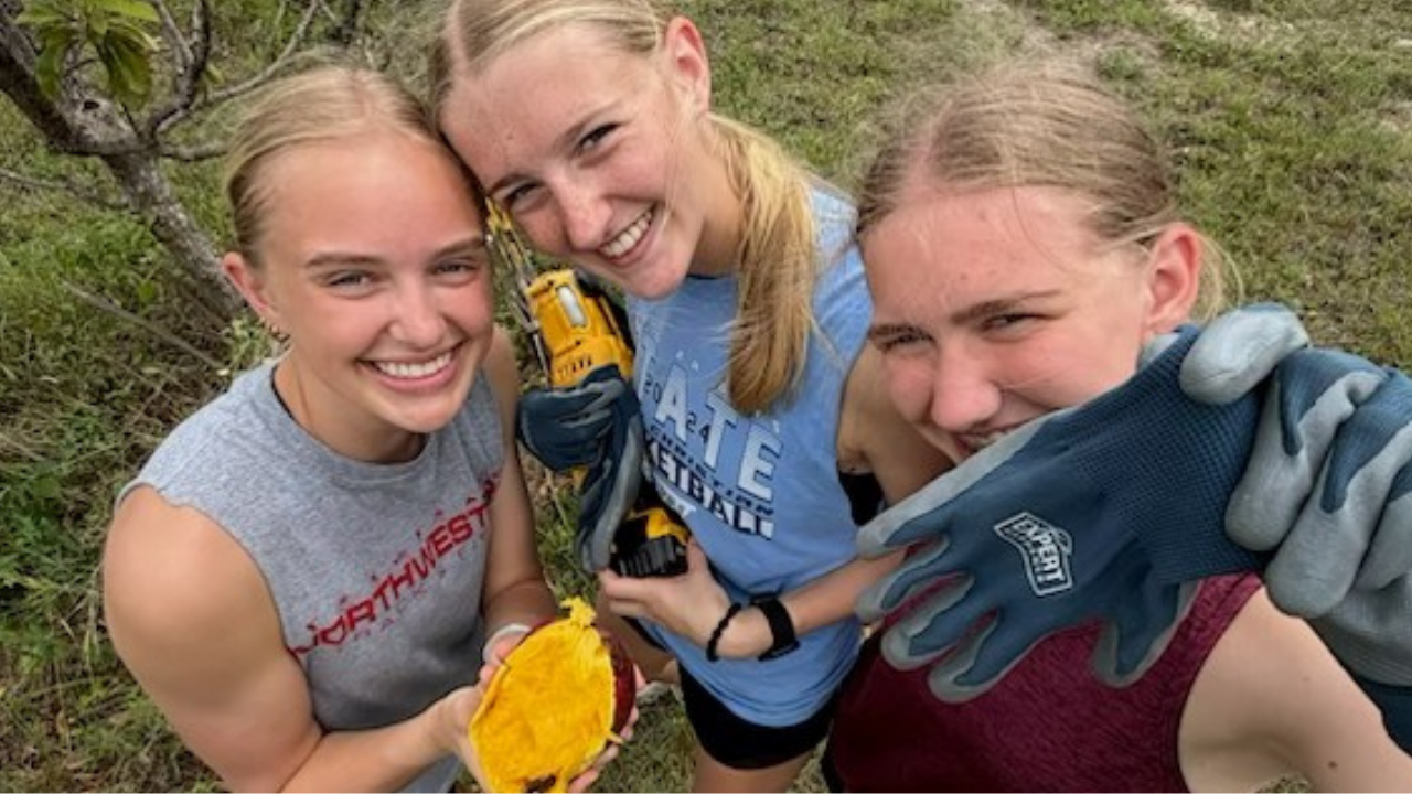Three female student volunteers from Sheldon, Iowa, smile together during a break from farm restoration work, experiencing the beauty of local agricultural immersion.
