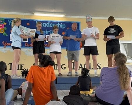 immanuel-reading-story Impact team volunteers stand on a school stage to read a story aloud to a group of local children gathered for an educational picnic.