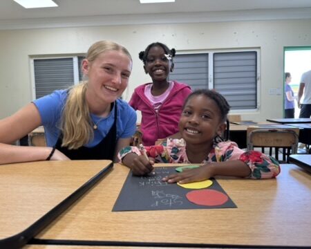 immanuel-smiling-at-desk A female youth volunteer smiles alongside local girls working on a classroom craft project during an intentional community mentorship day.