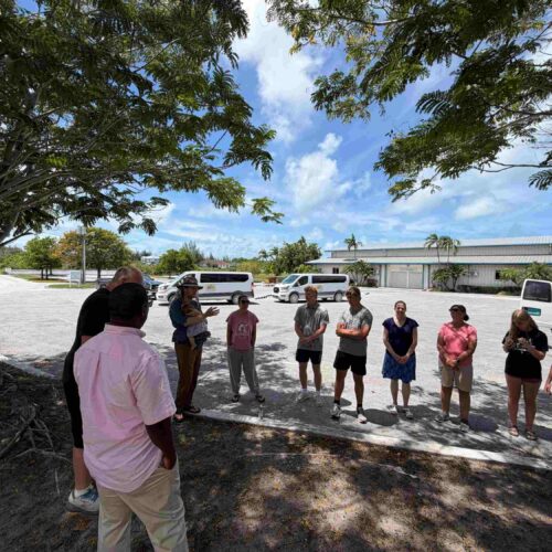 The complete Impact team gathers in a parking lot beneath tropical trees to pray before hand-delivering essential supply baskets to vulnerable neighborhood families.