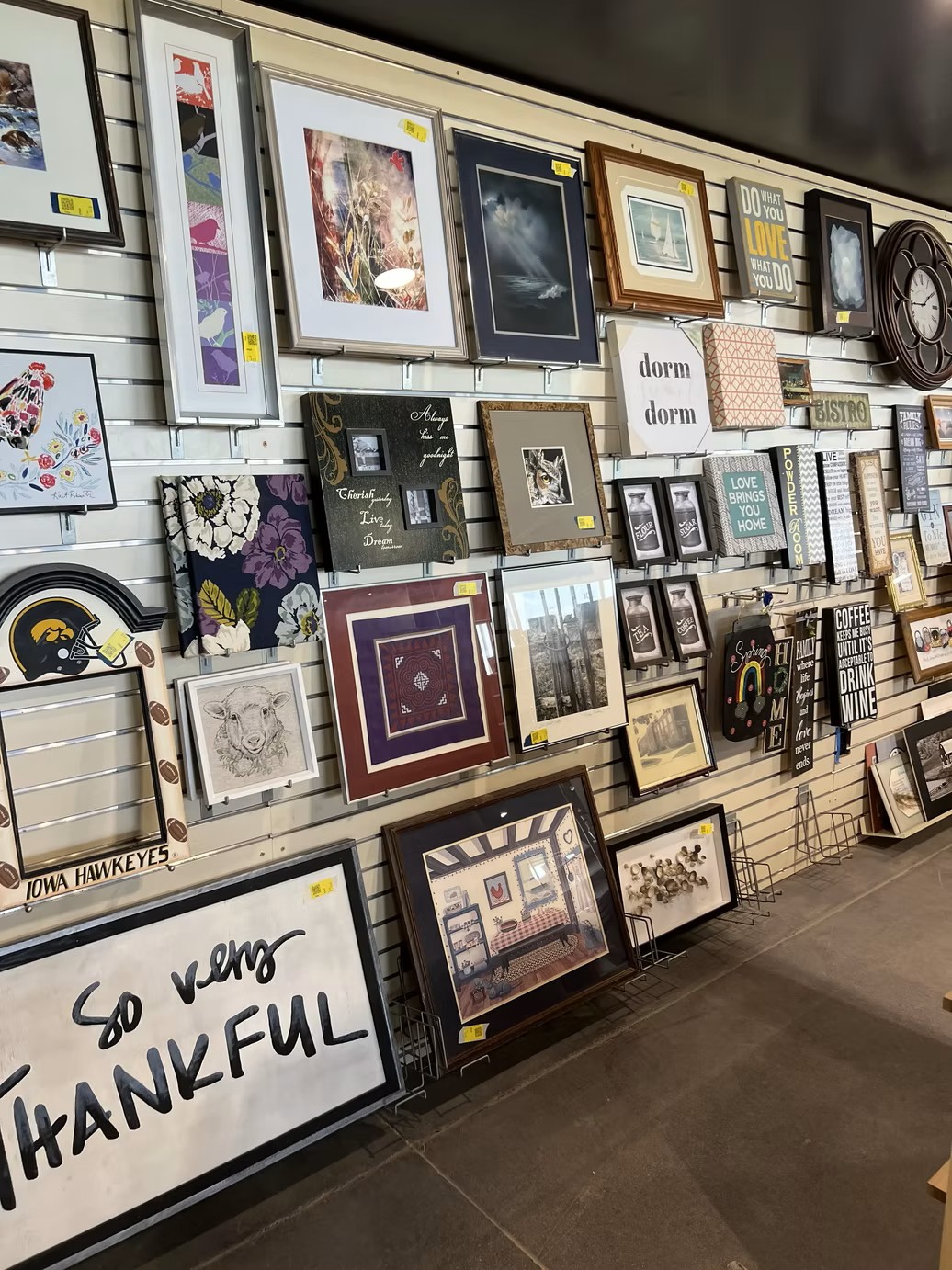 A slatwall display featuring a variety of framed wall art and signs at the new Jordan Creek Thrift Store.