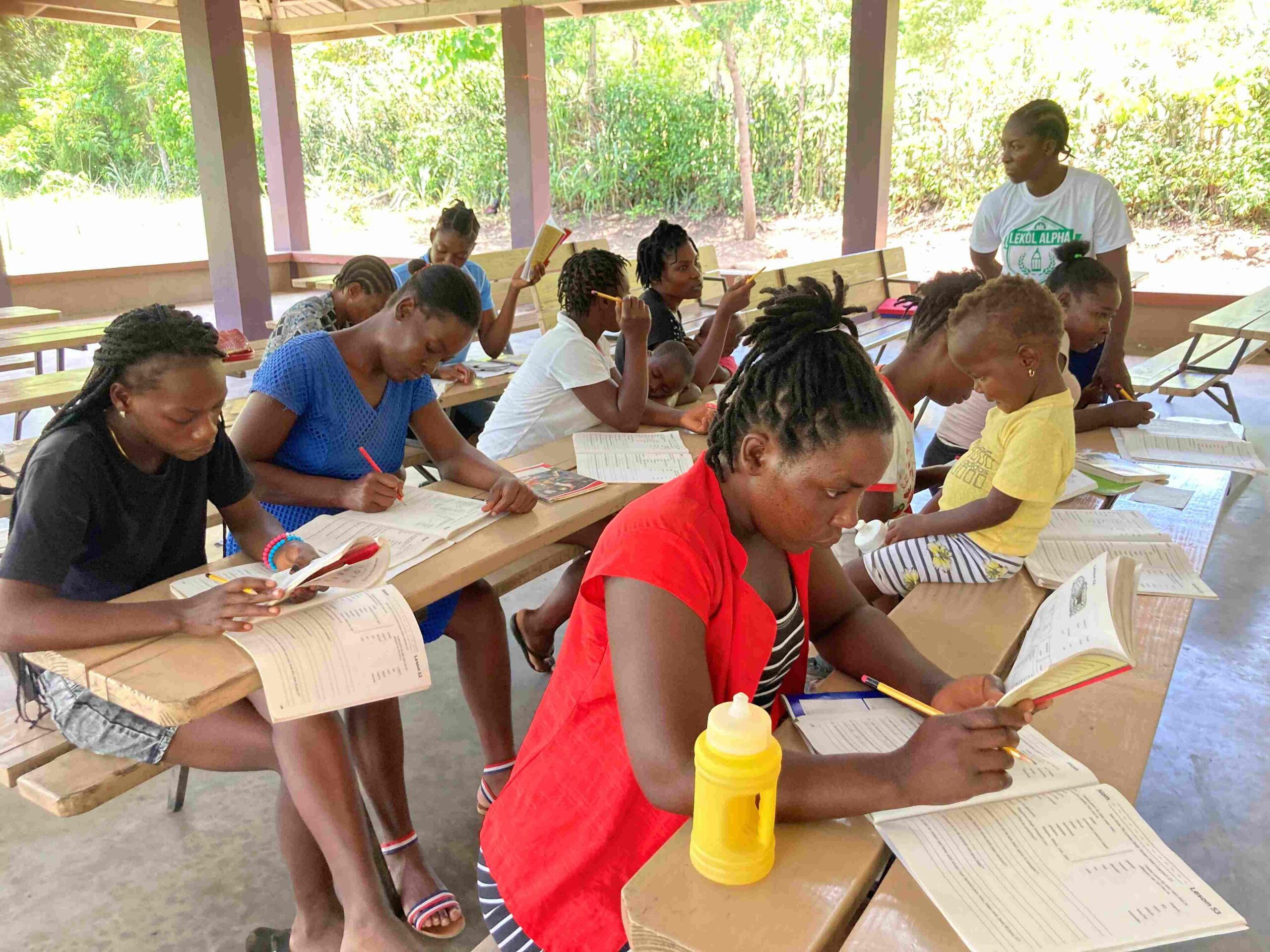 Haitian mothers sit together at wooden benches focused on reading workbooks and writing during an adult Lekòl Alpha class at Jean Boule.