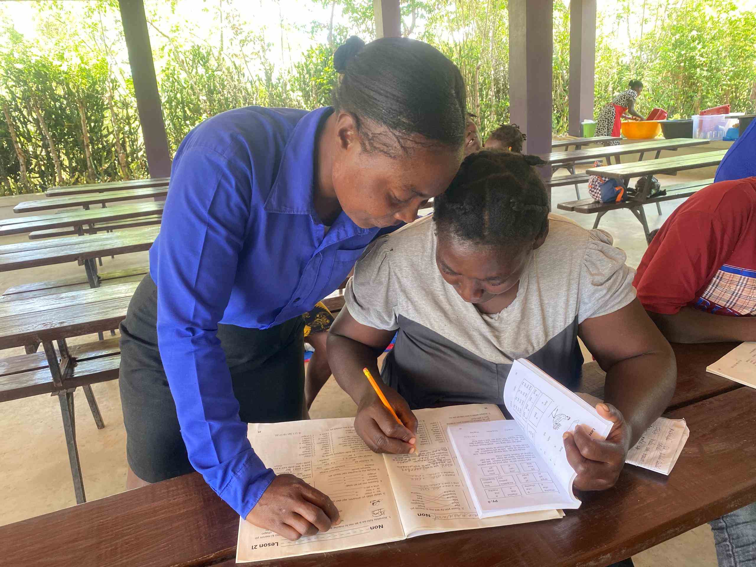 Lekòl Alpha teacher Monise leans over a table to guide a Haitian mother through a literacy workbook lesson at the Maliarette campus.